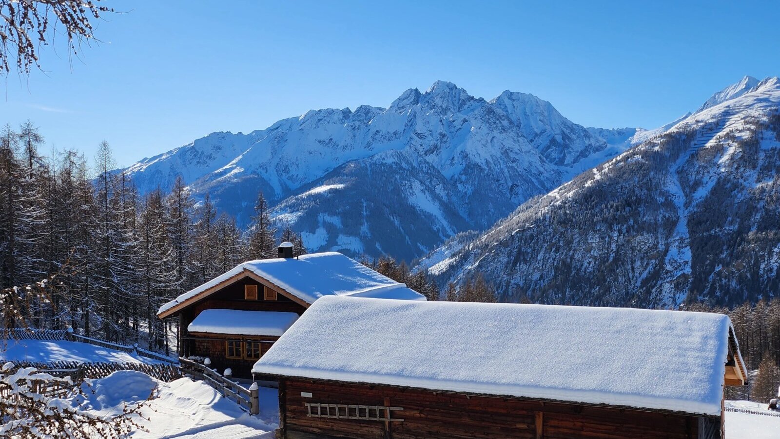 The snow-covered Almhütte and a second wooden building, surrounded by a winter landscape with snow-capped mountains.