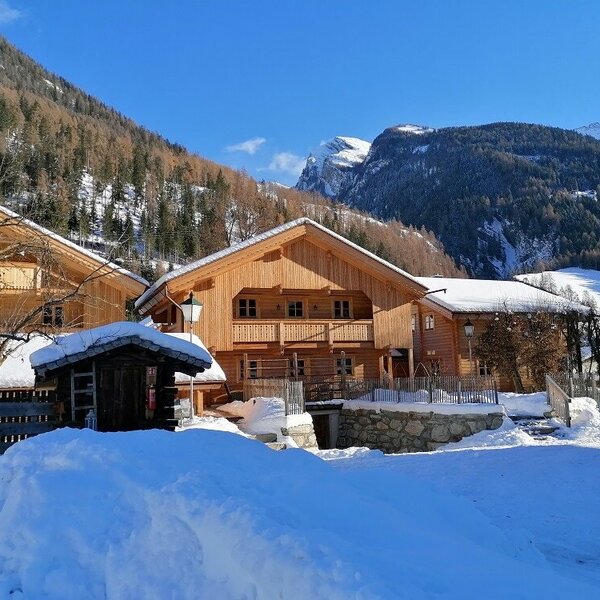 The wooden chalets of the Hotel & Chalets Schlosswirt, covered in snow and set against a backdrop of snow-covered mountains.