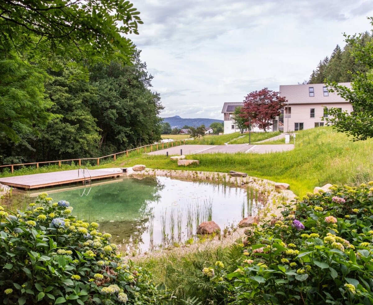 The natural pond features a wooden deck and is surrounded by green areas and flowers.
