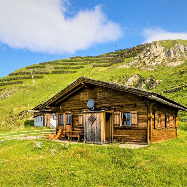 The Seppenalm Alpine Hut, a wooden structure situated on a grassy mountain slope, features an outdoor wooden bench and views of the surrounding landscape.