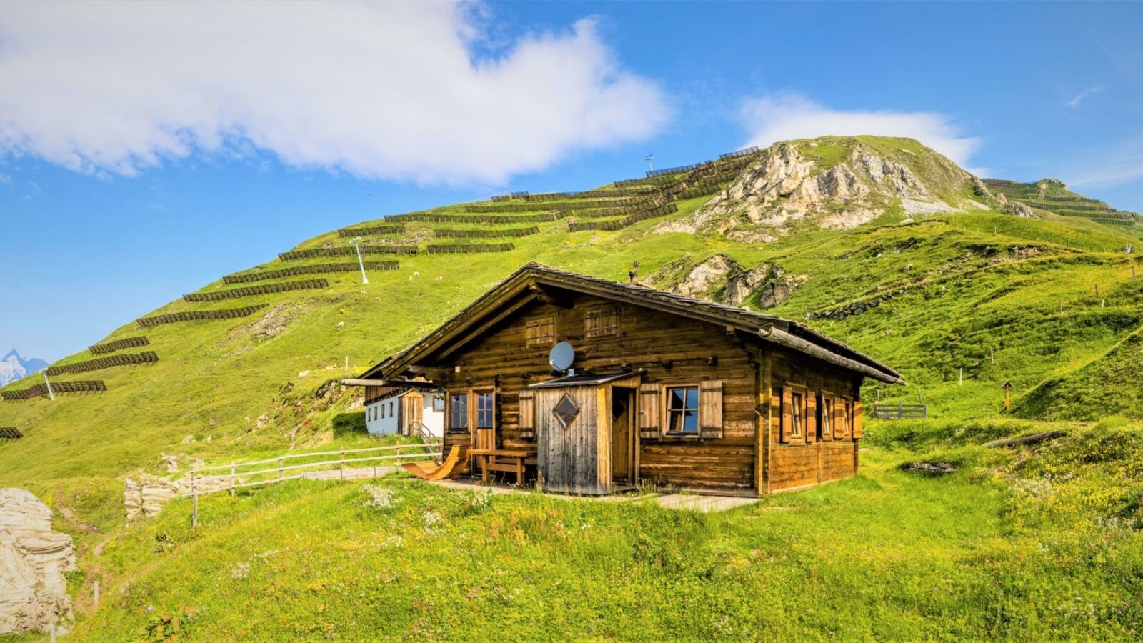 The Seppenalm Alpine Hut, a wooden structure situated on a grassy mountain slope, features an outdoor wooden bench and views of the surrounding landscape.