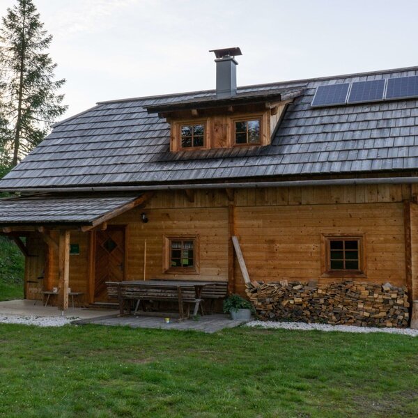 The Alpine Hut exterior, featuring a shingled roof with solar panels, a covered entrance, outdoor seating, and a stack of firewood.
