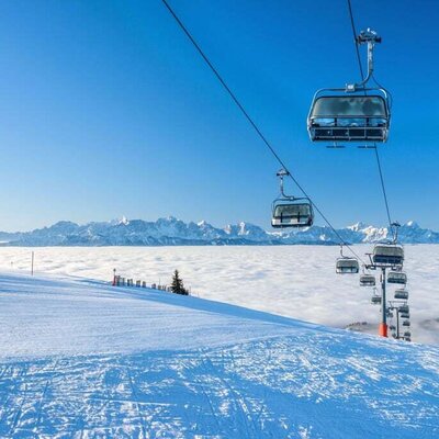 A ski slope with a chairlift and distant snow-capped mountains visible above a cloud layer, showcasing the winter sports environment surrounding the Farm House.