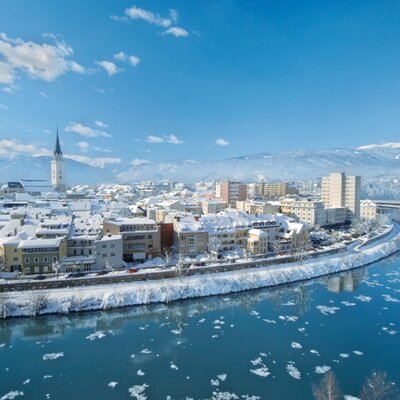 View of a snow-covered town featuring a church spire, a river with ice floes, and distant mountains, located near the Farm House.