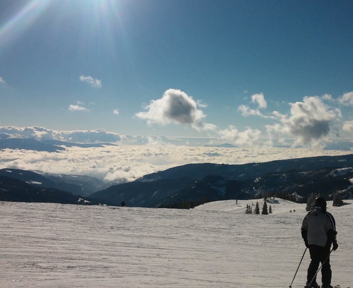A snow-covered ski slope with skiers and distant mountain peaks visible above a cloud inversion, characteristic of the Alpine Hut's setting.