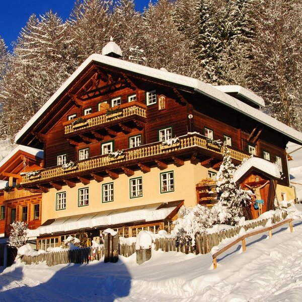 The farmhouse exterior features multiple wooden balconies and a snow-covered roof, set against a backdrop of snow-dusted trees.
