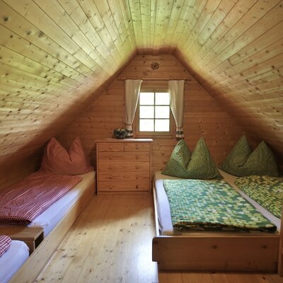 Upper floor bedroom in the Alpine Hut with two single beds, a wooden chest of drawers, and a window under a pitched wooden ceiling.