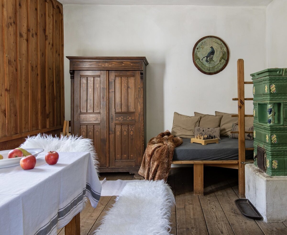 The Alpine Hut features a sleeping sofa, a traditional green tiled stove, and a wooden wardrobe, complemented by wooden wall paneling.