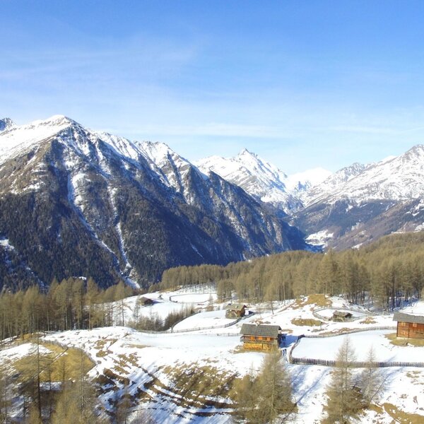 View of the snow-covered alpine landscape from the Alpine Hut, with snow-capped mountains, a valley, and scattered traditional wooden huts among bare larch trees. View of the snow-covered alpine landscape from the Alpine Hut, with snow-capped mountains, a valley, and scattered traditional wooden huts among bare larch trees.
