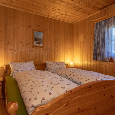 Double bedroom in the Alpine hut with natural wood paneling, a bed with floral bedding, and a window with checked and lace curtains.