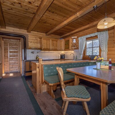 The alpine hut's kitchen and dining area features a wooden-paneled interior, equipped with wooden cabinets, a dining table with upholstered benches, and a television.