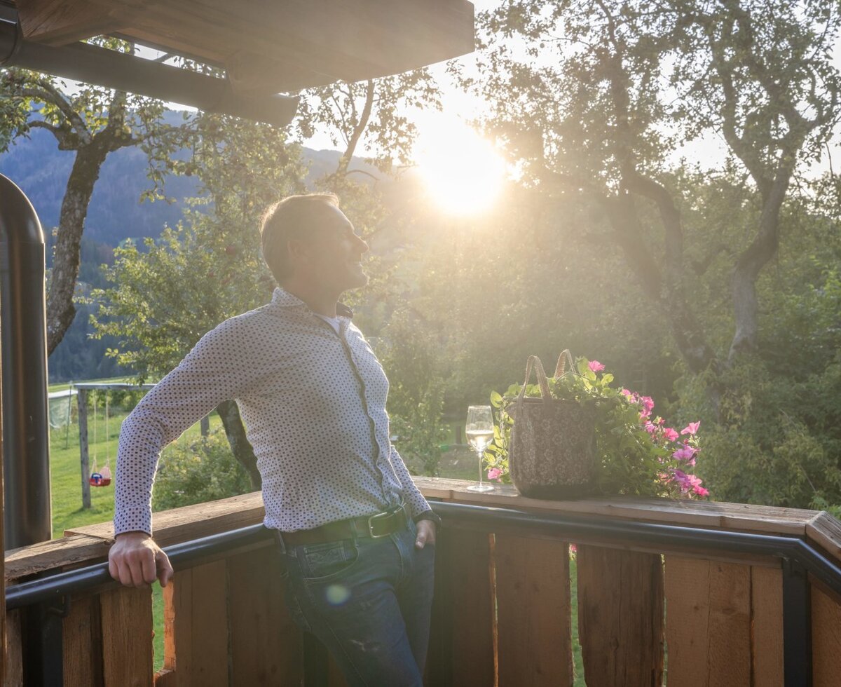 The chalet's wooden terrace, equipped with a glass of white wine and potted flowers, offers views of the surrounding mountains and trees in bright sunlight.