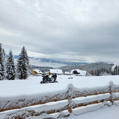A winter view of the snow-covered landscape surrounding the Alpine Hut, with evergreen trees, nearby farm buildings, and the Lavanttal valley in the background.