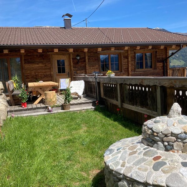 The Alm features a wooden terrace with a table and chairs, surrounded by a green lawn and mountain views.