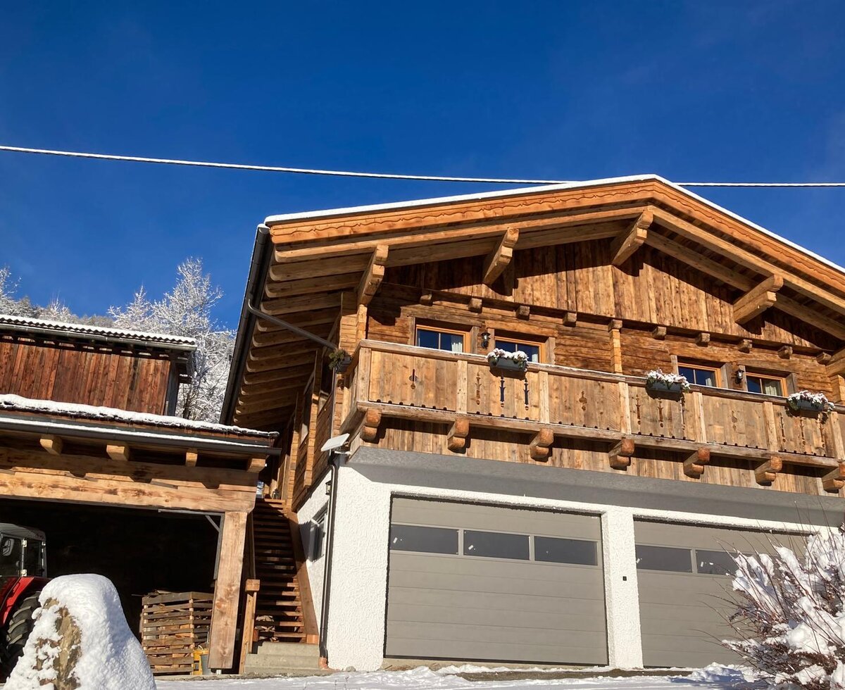 Exterior of the Alm with a wooden balcony, windows, two garage doors, and an external staircase, set in a snowy landscape.
