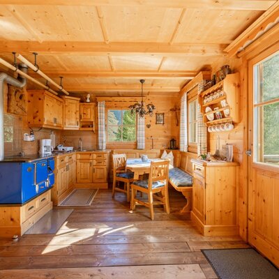 The Alm's kitchen with a wood-burning stove, corner bench, dining table, and windows offering a view of greenery.