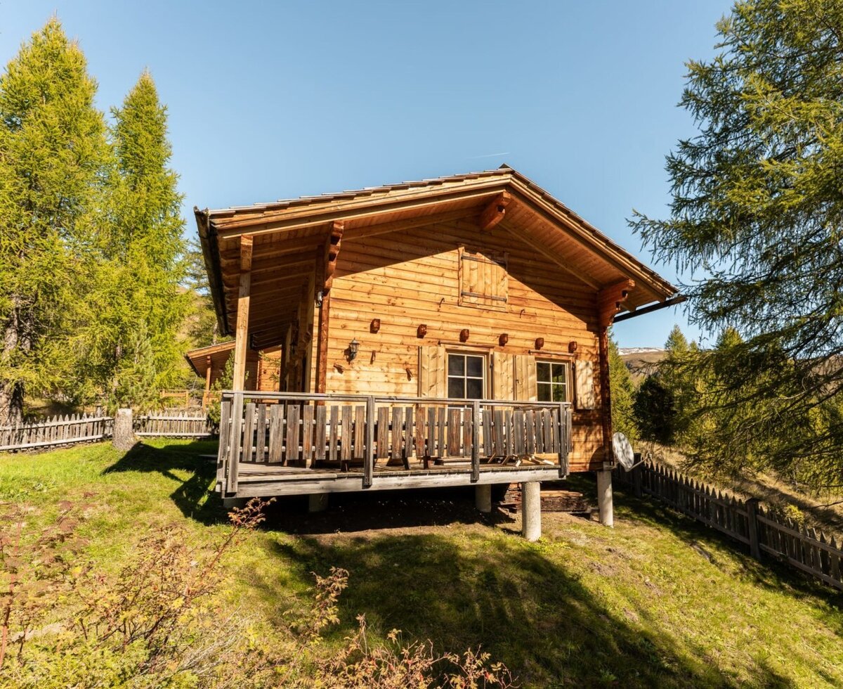 The Alm from the outside, featuring a wooden facade, balcony, and windows with shutters, nestled in a green landscape with trees and a wooden fence.