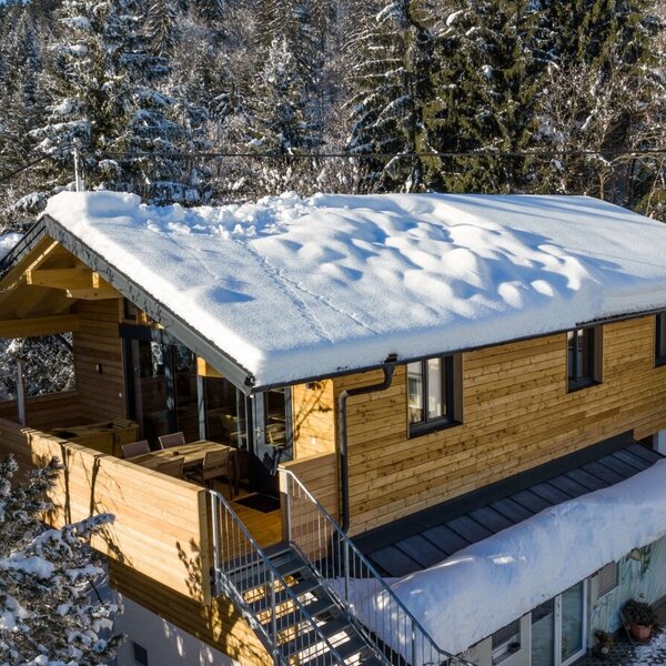 The farm house features a wooden exterior, a snow-covered roof, and a balcony with an outdoor dining area, surrounded by snowy forests.