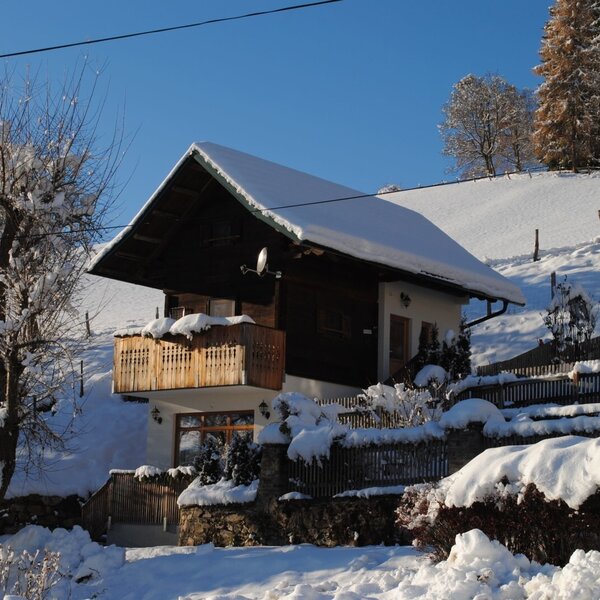 The vacation rental with a snow-covered roof and wooden balcony, located on a sunny, snow-covered hillside.