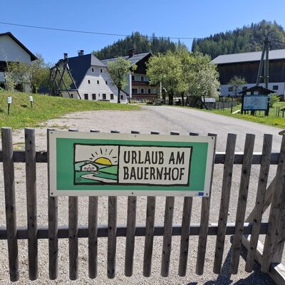 The entrance to the Farm House, identified by a wooden gate and an "Urlaub am Bauernhof" sign, with farm buildings and a children's slide in the background.