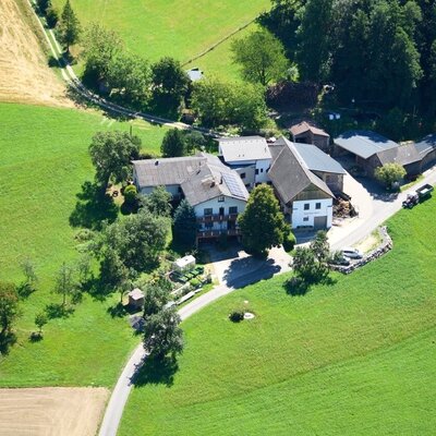 Aerial view of the farmhouse with its buildings, surrounded by green meadows and trees, with an access road.
