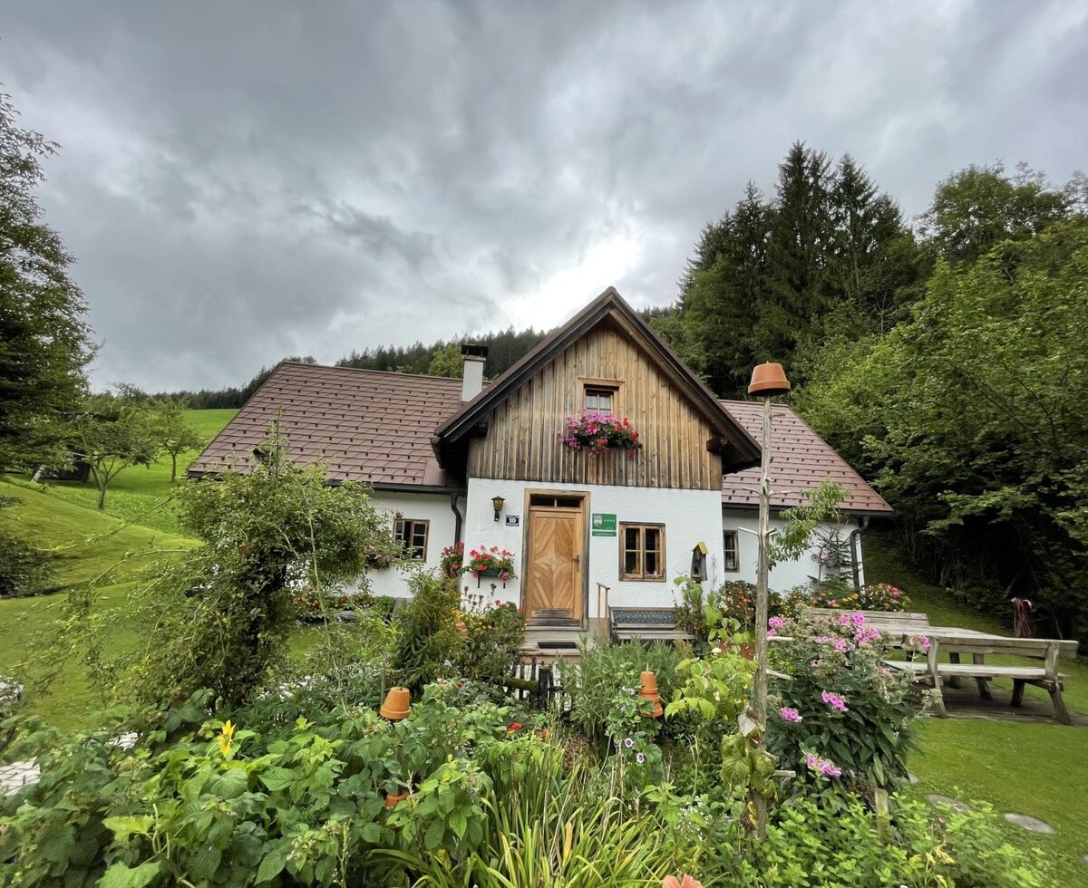 The Farm House exterior, featuring a traditional design, a surrounding garden, and an outdoor picnic table on the lawn, with hills and forest in the background.
