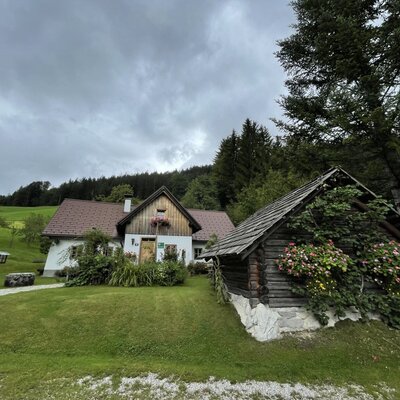 The exterior of the Farm House, featuring a white facade, wooden gables, and a log cabin outbuilding, surrounded by a green lawn and forest.