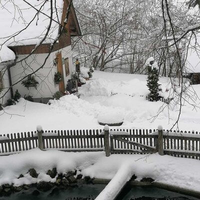 The farm house exterior covered in deep snow, featuring snow-covered trees, a wooden fence, and a pond with a snow-laden wooden bridge.