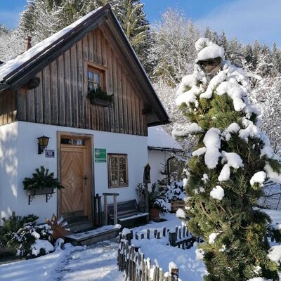 The snow-covered farmhouse exterior with its wooden upper facade and white lower walls, featuring a path to the entrance and surrounding winter trees.