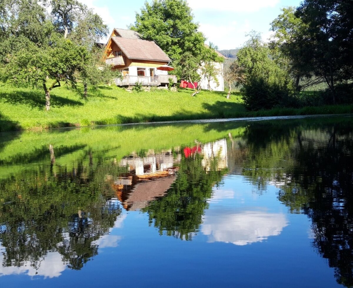 The Bauernhof's Knusperhaus by the pond, surrounded by greenery, with its reflection in the water.