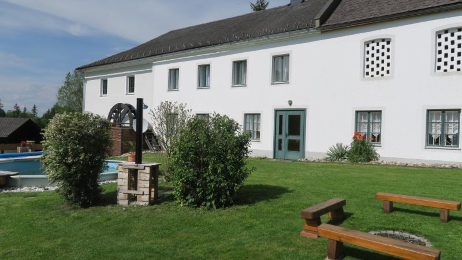The farm house exterior features a green lawn with wooden benches surrounding a fire pit, a water wheel, and a swimming pool.
