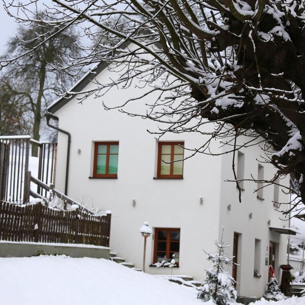 The white farmhouse exterior, covered in snow, includes a fenced terrace area and a large snow-covered tree. The white farmhouse exterior, covered in snow, includes a fenced terrace area and a large snow-covered tree.