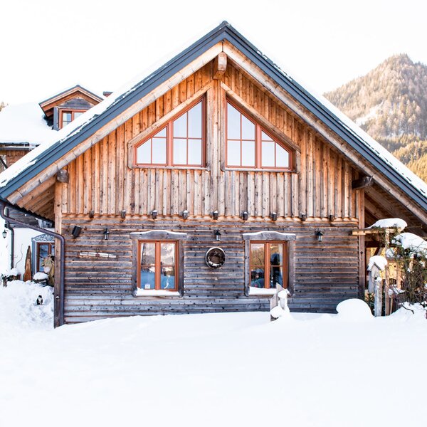 The exterior of the wooden farmhouse, featuring snow-covered grounds and a mountain backdrop.