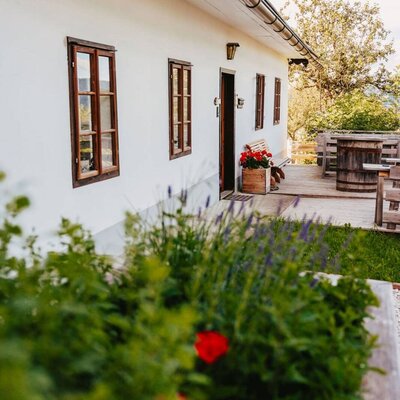 The farmhouse exterior features a wooden deck with a bench and barrel table, adjacent to garden beds with lavender and red flowers.
