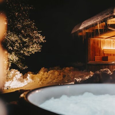The farmhouse features an outdoor hot tub and a wooden sauna hut, surrounded by snow and illuminated at night.