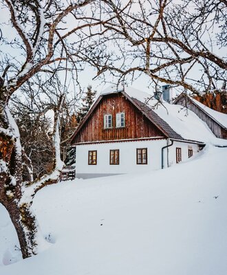 The farmhouse exterior, featuring its wooden upper facade and white lower walls, surrounded by snow and bare trees in winter.