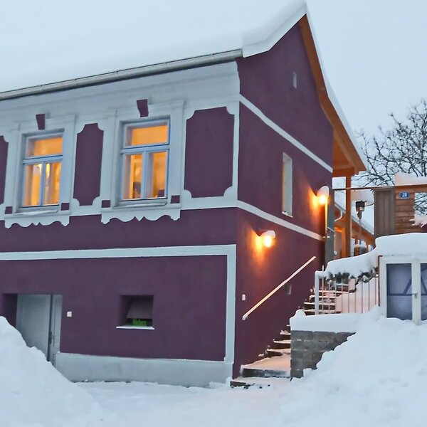 The farmhouse exterior of Traubengarten-Gaestehaus, featuring its purple facade, white trim, and illuminated windows and outdoor staircase, covered in snow.