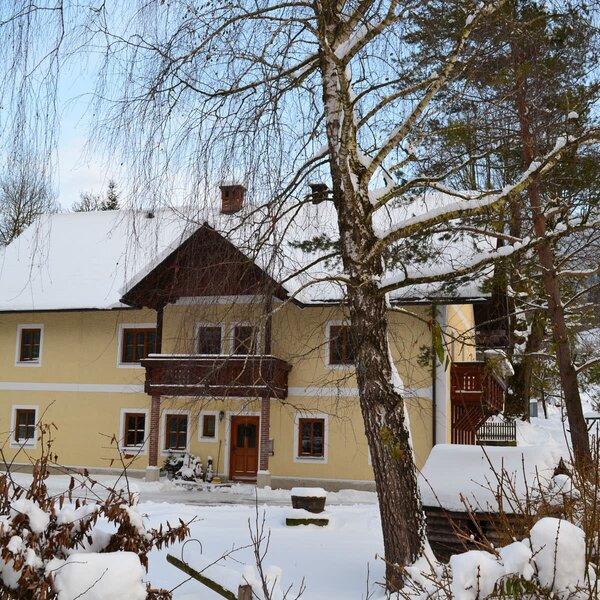 The exterior of the Farm House, a yellow building with a snow-covered roof and a wooden balcony, surrounded by winter trees.