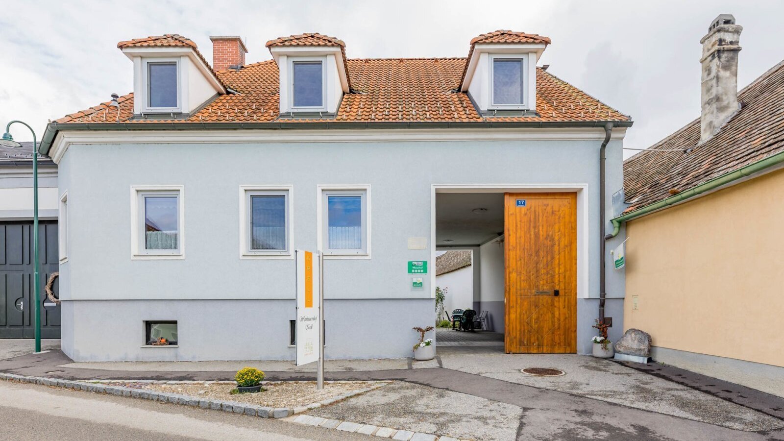 Exterior view of the house, featuring a light blue facade, red tiled roof, and a large wooden gate.