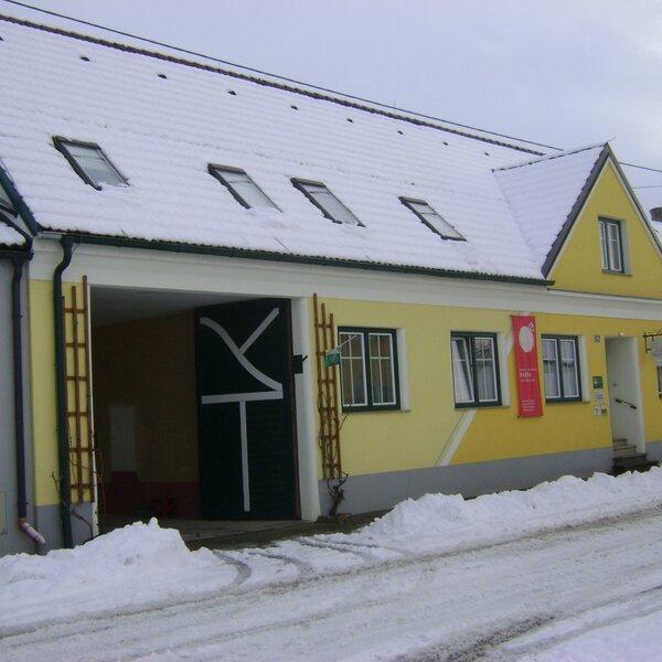 The yellow and gray exterior of the farmhouse, featuring a snow-covered roof with skylights and a large open entrance on the left.