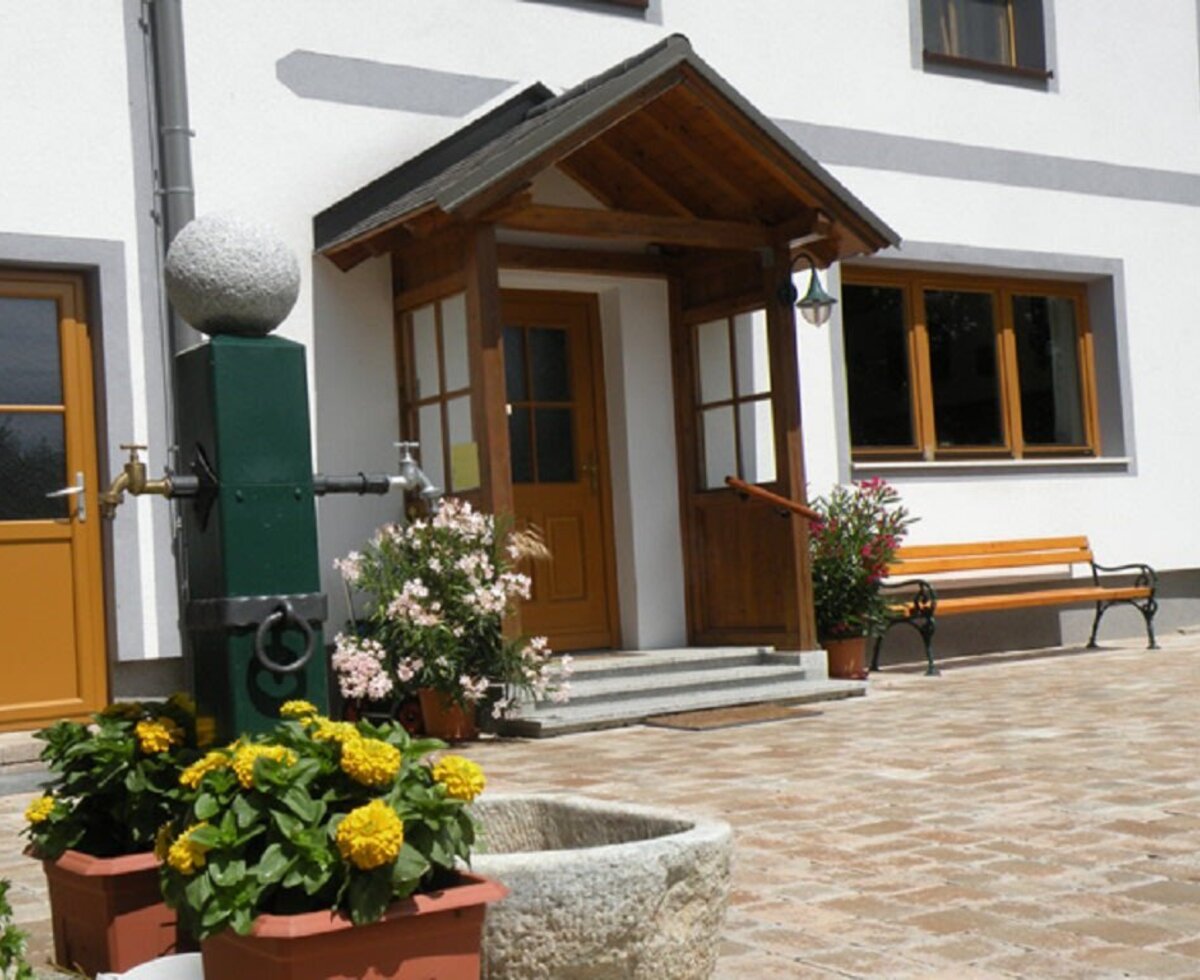 The entrance area of the farm house, featuring a covered wooden porch, a water pump, and a wooden bench.