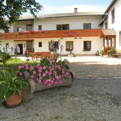 The farmhouse exterior features a wooden balcony, an outdoor seating area with tables and benches, and a paved courtyard with floral decorations.
