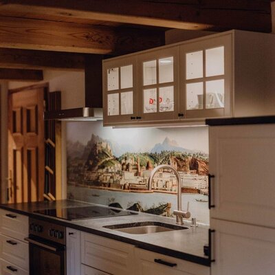 The farmhouse kitchen is equipped with white cabinets, a dark countertop, a sink, an integrated oven and hob, illuminated upper cabinets, and a landscape mural backsplash.