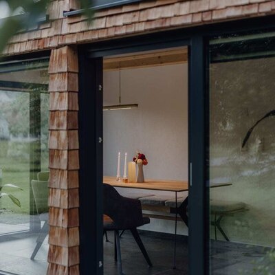 The dining area in the chalet with a wooden table, chairs, and a bench, visible through the open glass front.