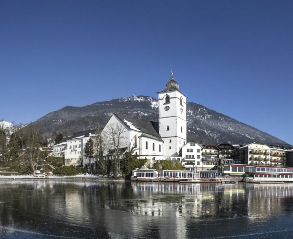 Lake Wolfgangsee with the village, its pilgrimage church, and snow-capped mountains, showcasing the scenic surroundings of the farmhouse.