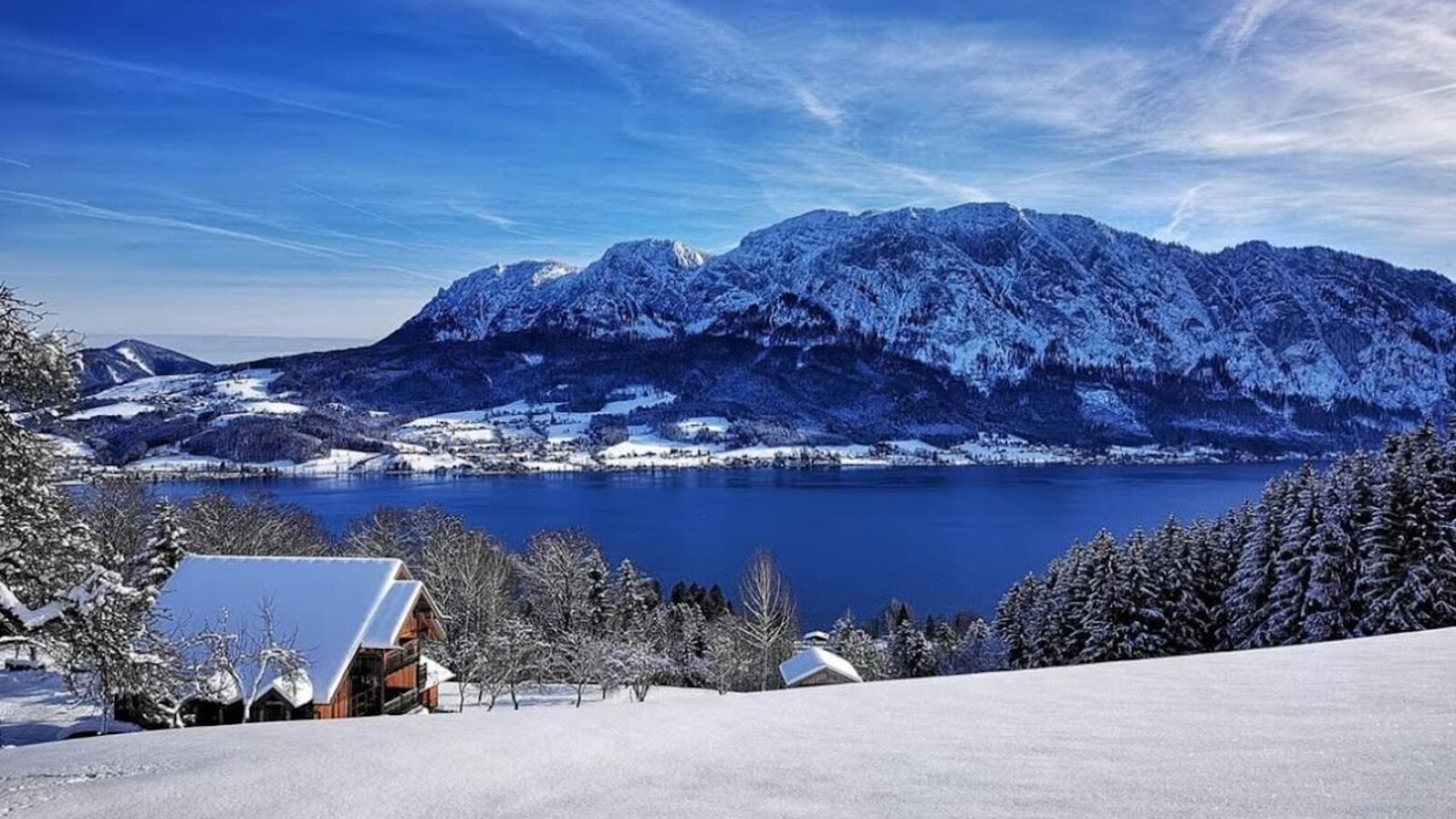 Winter landscape featuring a farmhouse, Lake Attersee, and the snow-covered Höllengebirge mountains.