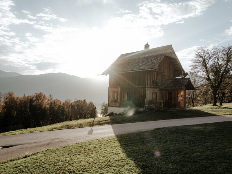 The wooden farmhouse exterior, set on a grassy hill with mountains in the background and a road leading to the property.