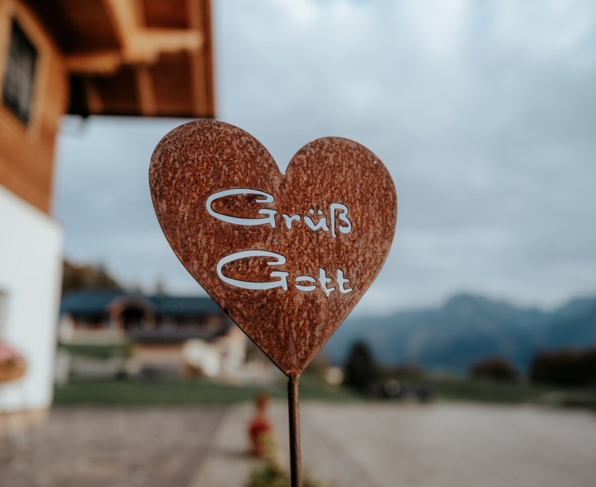 A rusty heart-shaped sign displaying "Grüß Gott" welcomes guests to the Farm House, with mountains visible in the background.