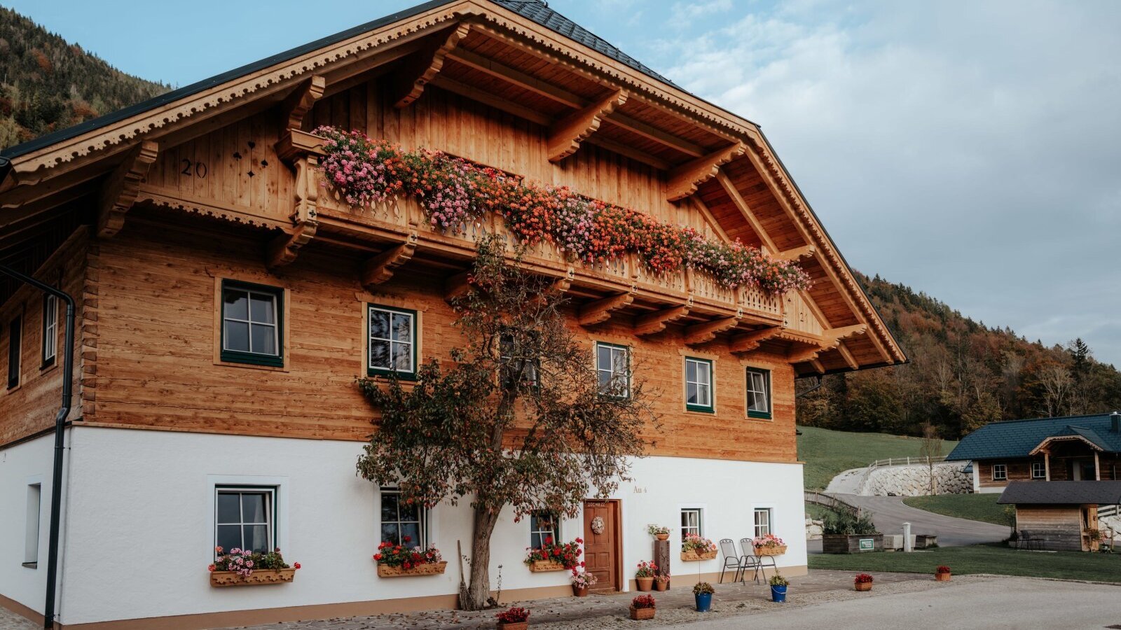 The traditional Farm House exterior features a wooden upper floor with flower-filled balconies, a white lower level, and an entrance with outdoor seating.
