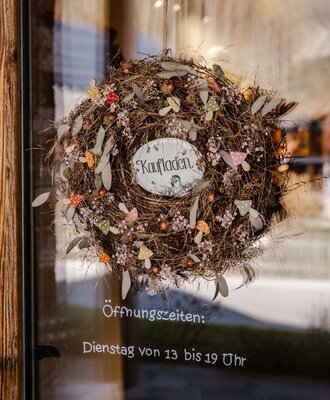 Entrance to the Bauernhof's shop, featuring a decorative wreath and opening hours on the glass door.